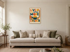 Live In A Little More Colour — A2 artwork shown in a room with a natural wood frame; select a framed finish to include a frame